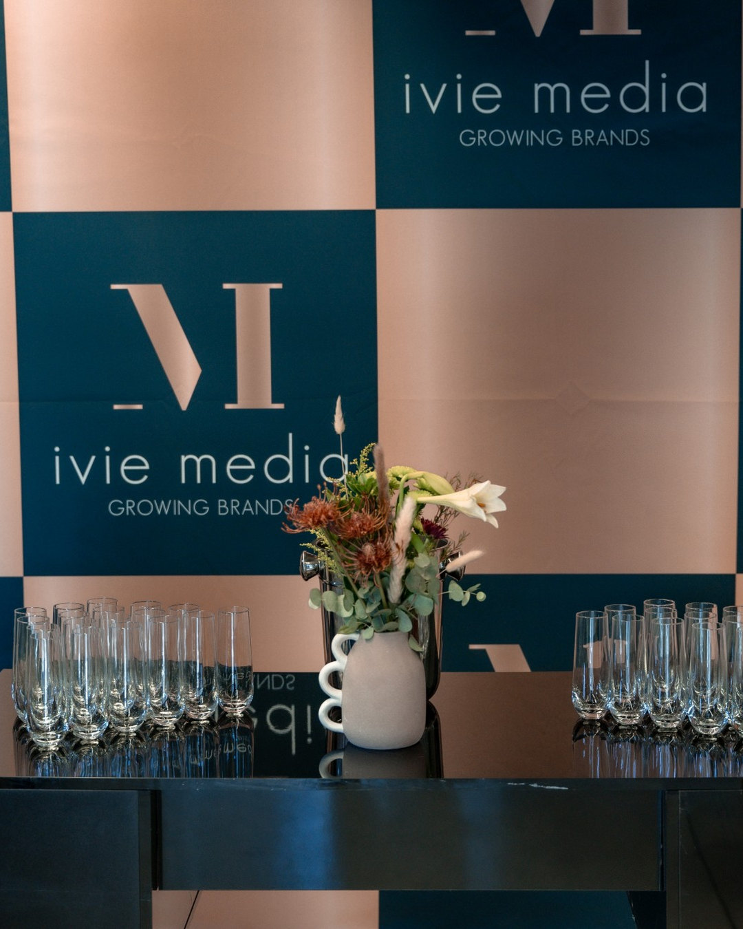 An Ivie Media branded backdrop with a flower arrangement and glasses on a drinks table